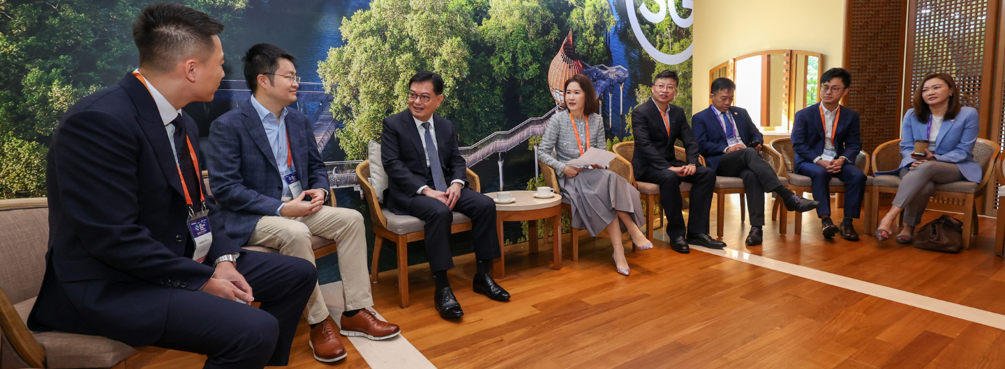 Nine people sit in a circle, business attire, in front of a backdrop of nature.